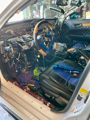 The interior of a 2013 Lexus GS450h mid-retrofit, dashboard removed, wiring looms exposed across the footwell and seat, with tools and control modules laid out